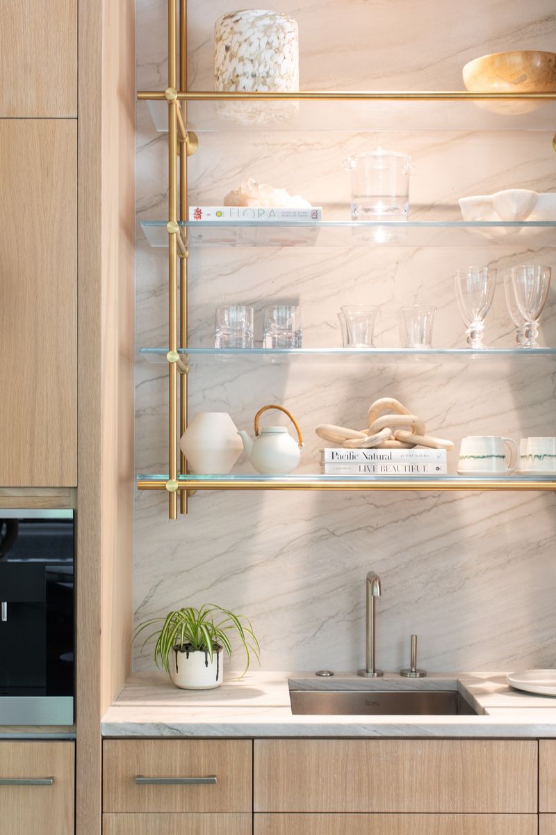 Modern kitchen with collector's series ceiling mounted 1 bay warm brass shelving unit, hanging with 4 glass shelves by Amuneal. Kitchen bar sink decorated with minimalist home style pieces with light tan oak cabinets , crystal clear drinking glasses and light pastel kitchenware.
